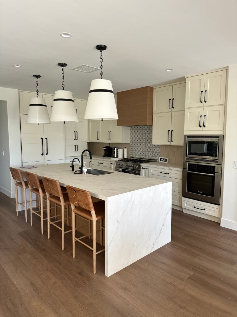 Modern kitchen featuring a large island with a white marble countertop, wooden bar stools, and stylish pendant lighting. The kitchen includes beige cabinets, a gas stove, and a patterned backsplash.